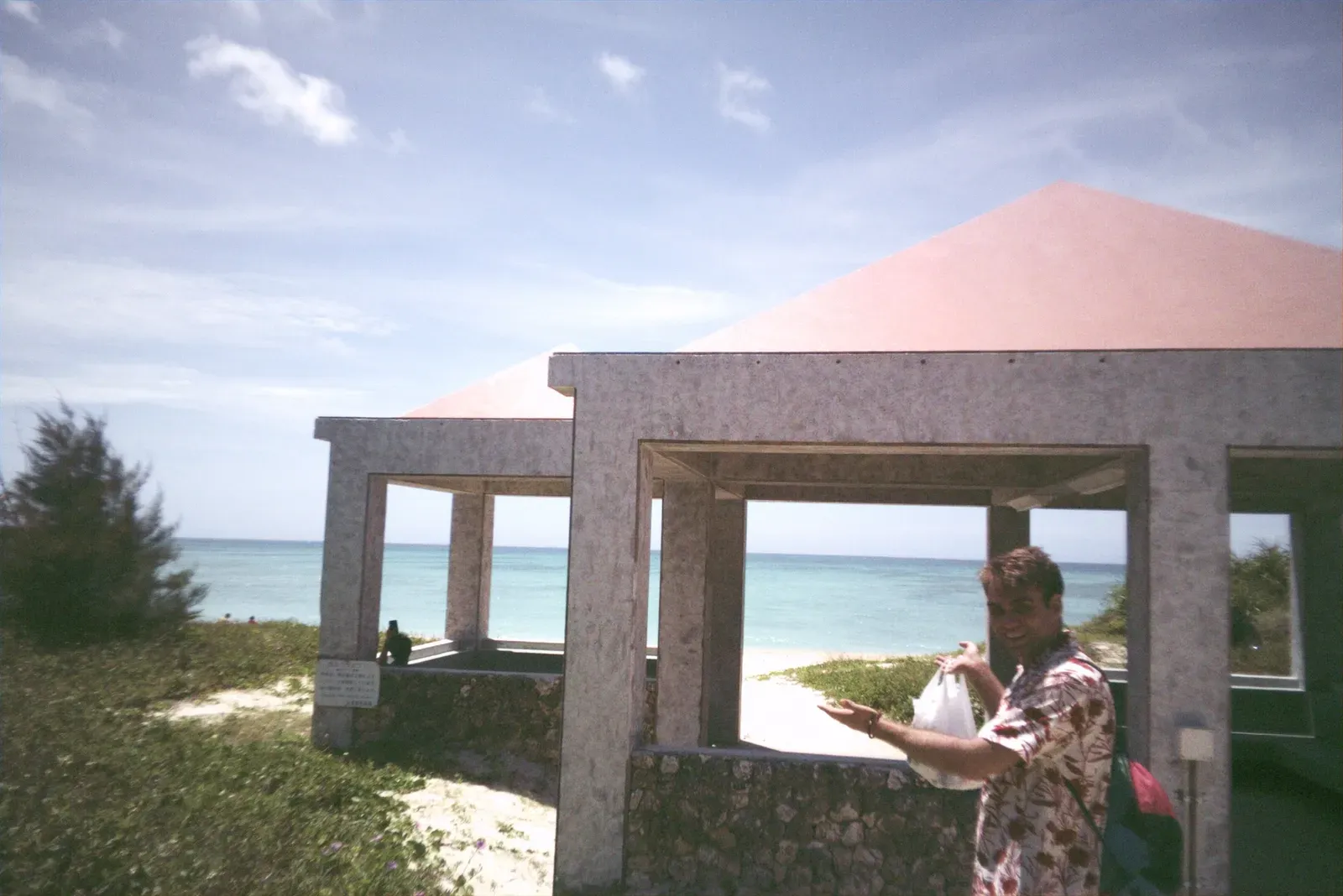 Itay grinning in a floral shirt beside a pink-roofed beach shelter, turquoise sea behind